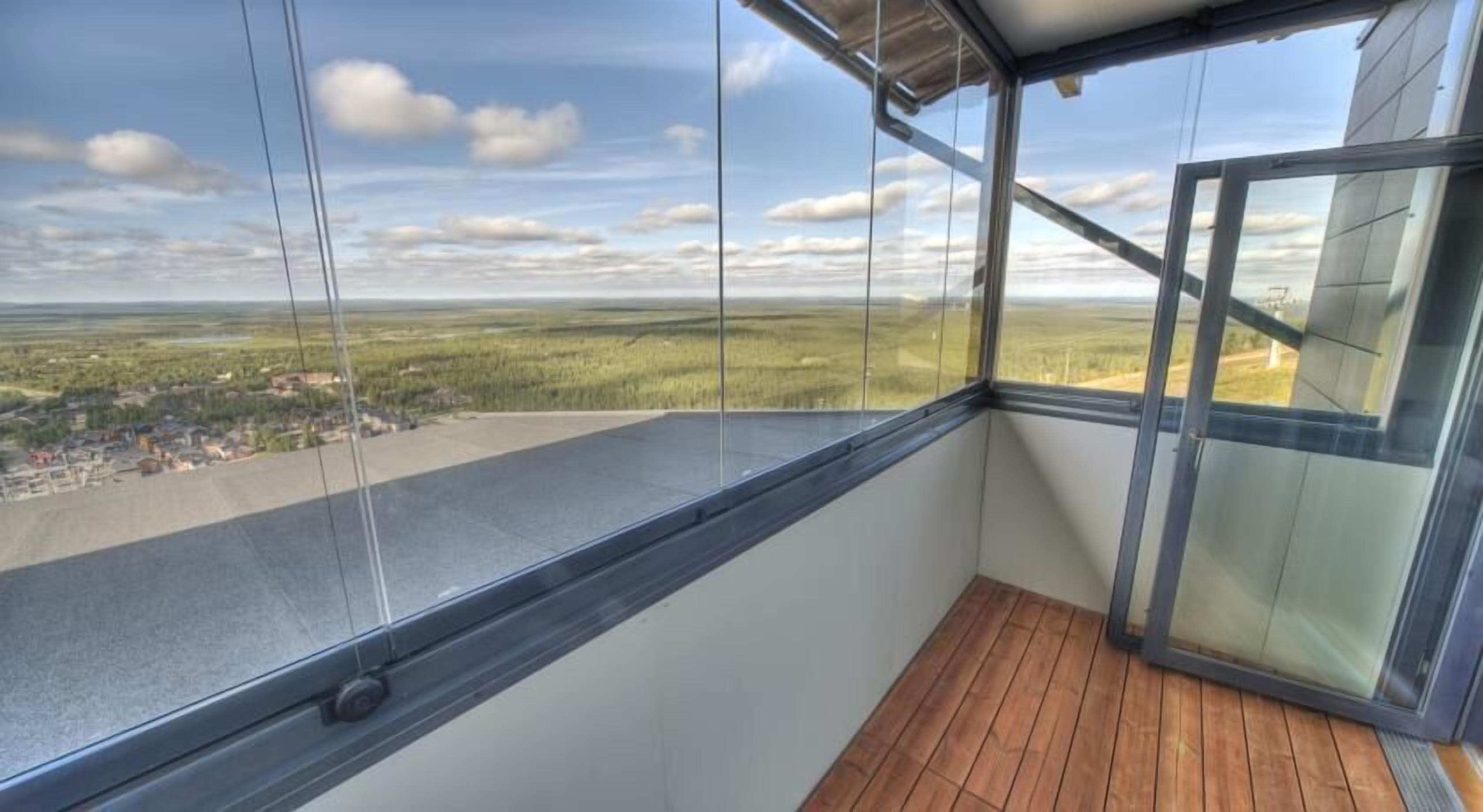 A balcony with a wooden floor and a glass wall offering a view of a rural landscape.