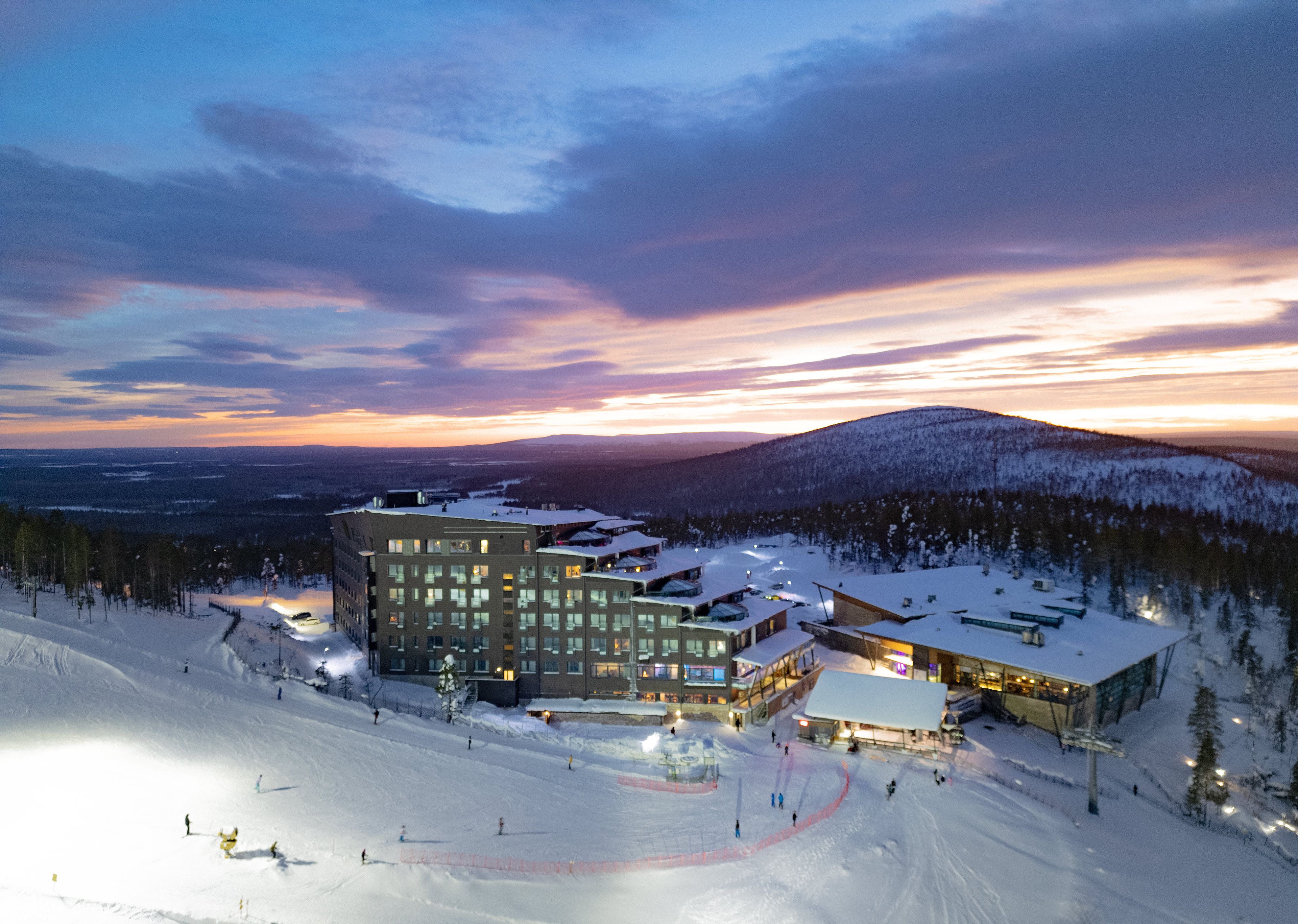 A snow-covered ski resort with a modern building complex, ski slopes, and a mountainous backdrop.