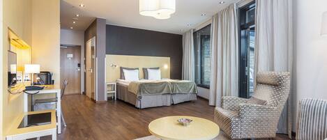 Una moderna camera d'hotel con un letto, un'area salotto con una poltrona e un tavolino, una scrivania con una sedia e un'ampia finestra con tende.