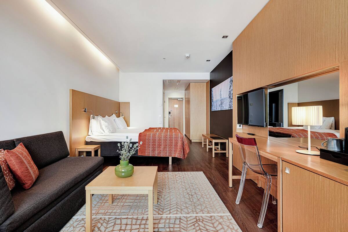 Una habitación de hotel moderna con sofá, cama con cubrecama estampado, escritorio con silla, televisión y un espejo grande.