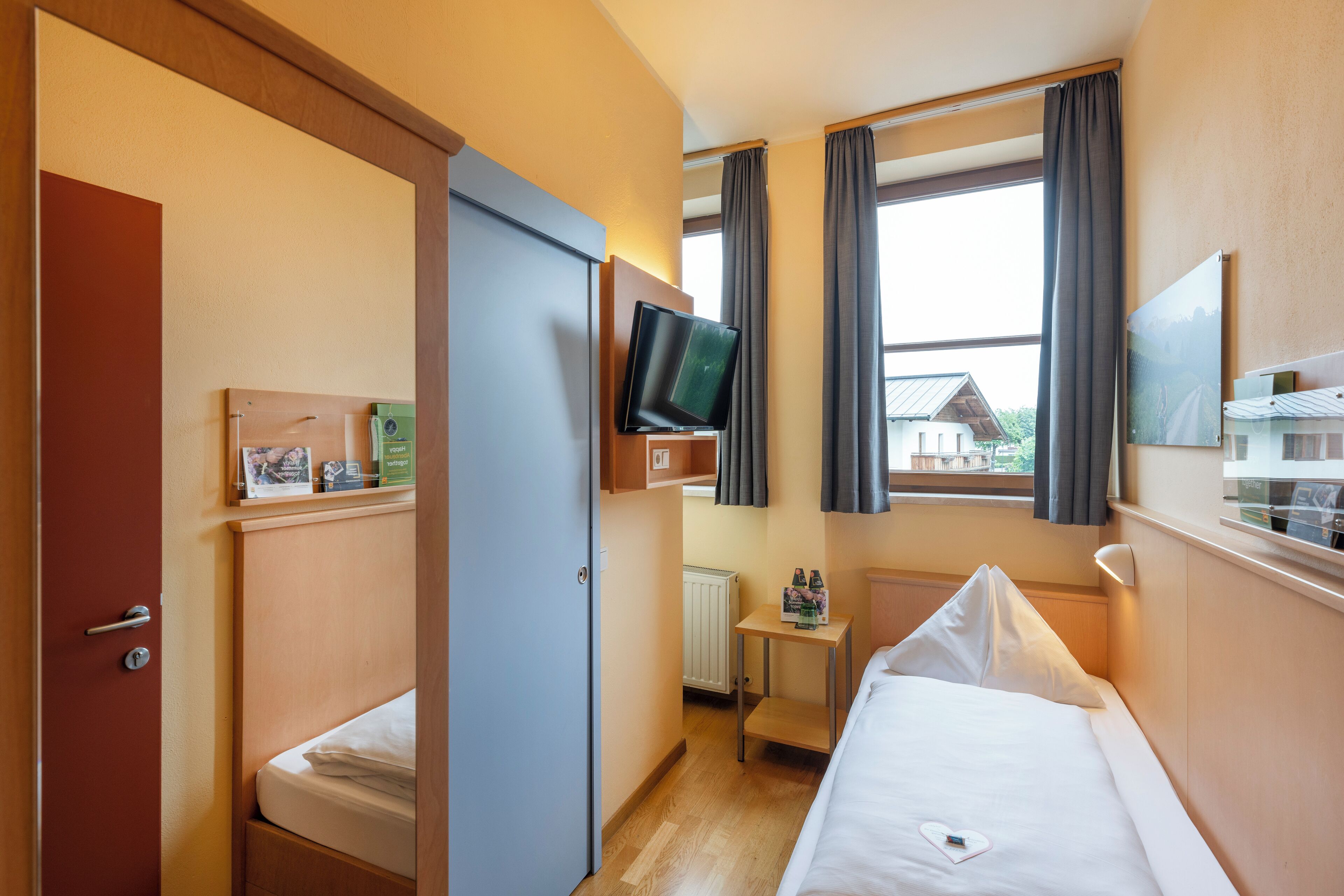 Une chambre d’hôtel avec un lit, une petite table, un téléviseur fixé au mur et une fenêtre avec des rideaux.