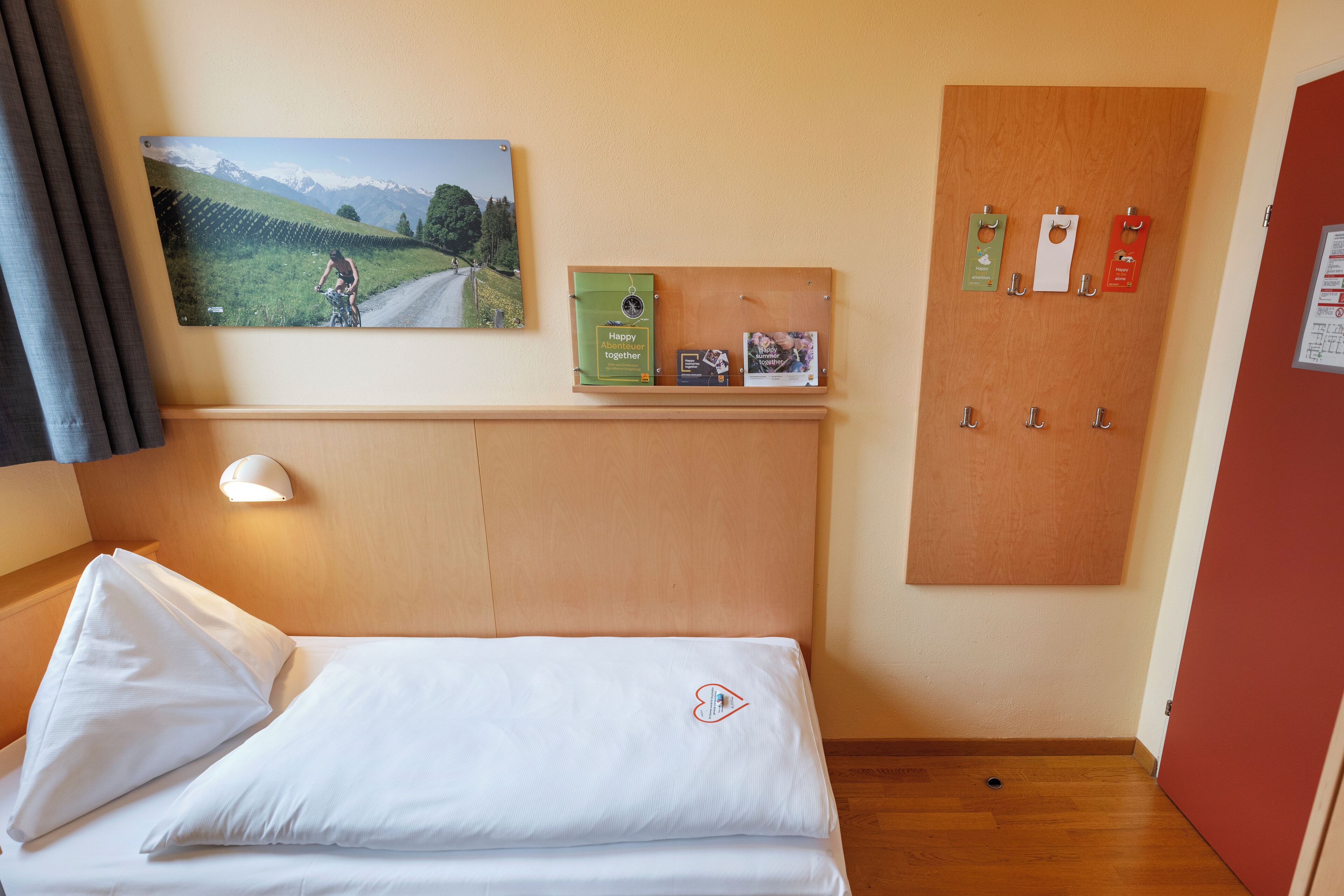 Habitación de hotel con una cama, un cuadro colgado de un ciclista en una carretera, un tablón de anuncios con tarjetas y una puerta de madera.