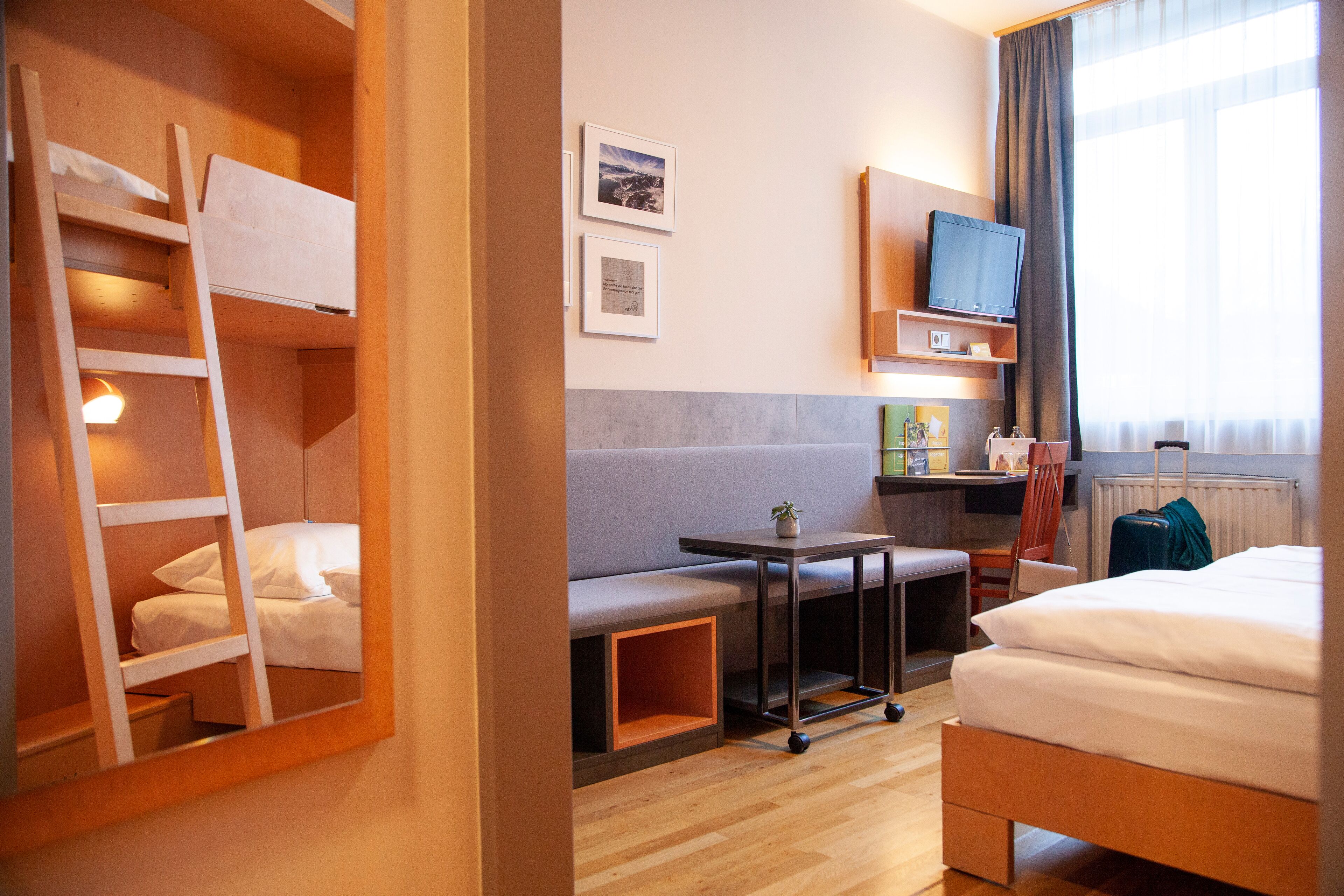 A hotel room with a bunk bed, a desk with a chair, a small table, a TV, and a window with curtains.