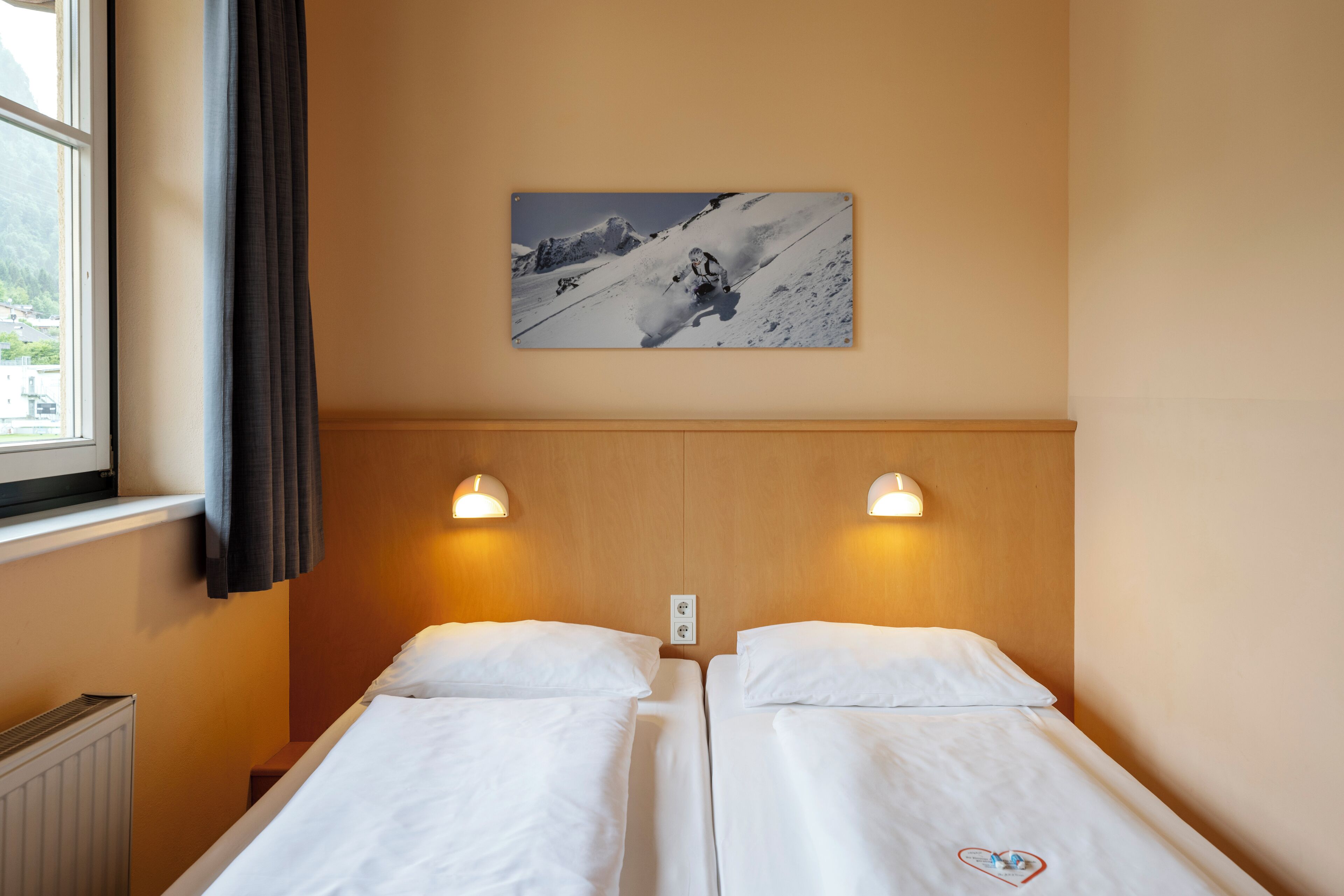 Una habitación de hotel con dos camas individuales, una ventana con cortinas, un cuadro colgado en la pared de un esquiador y dos luces de pared.