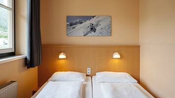 Una habitación de hotel con dos camas individuales, una ventana con cortinas, un cuadro colgado en la pared de un esquiador y dos luces de pared.
