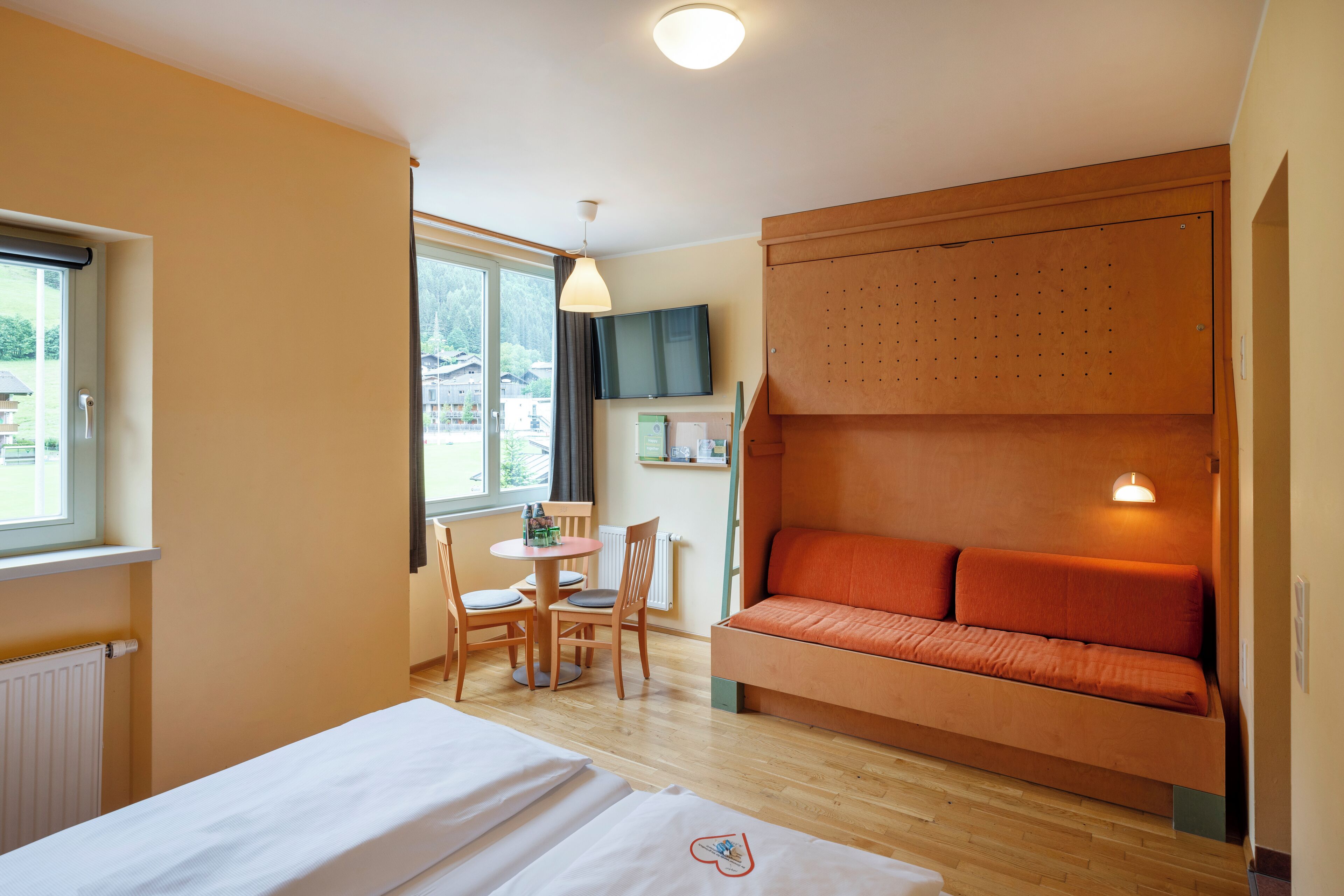 A hotel room with a bed, a wooden sofa, a dining table with chairs, a TV, and a window with a view of houses.