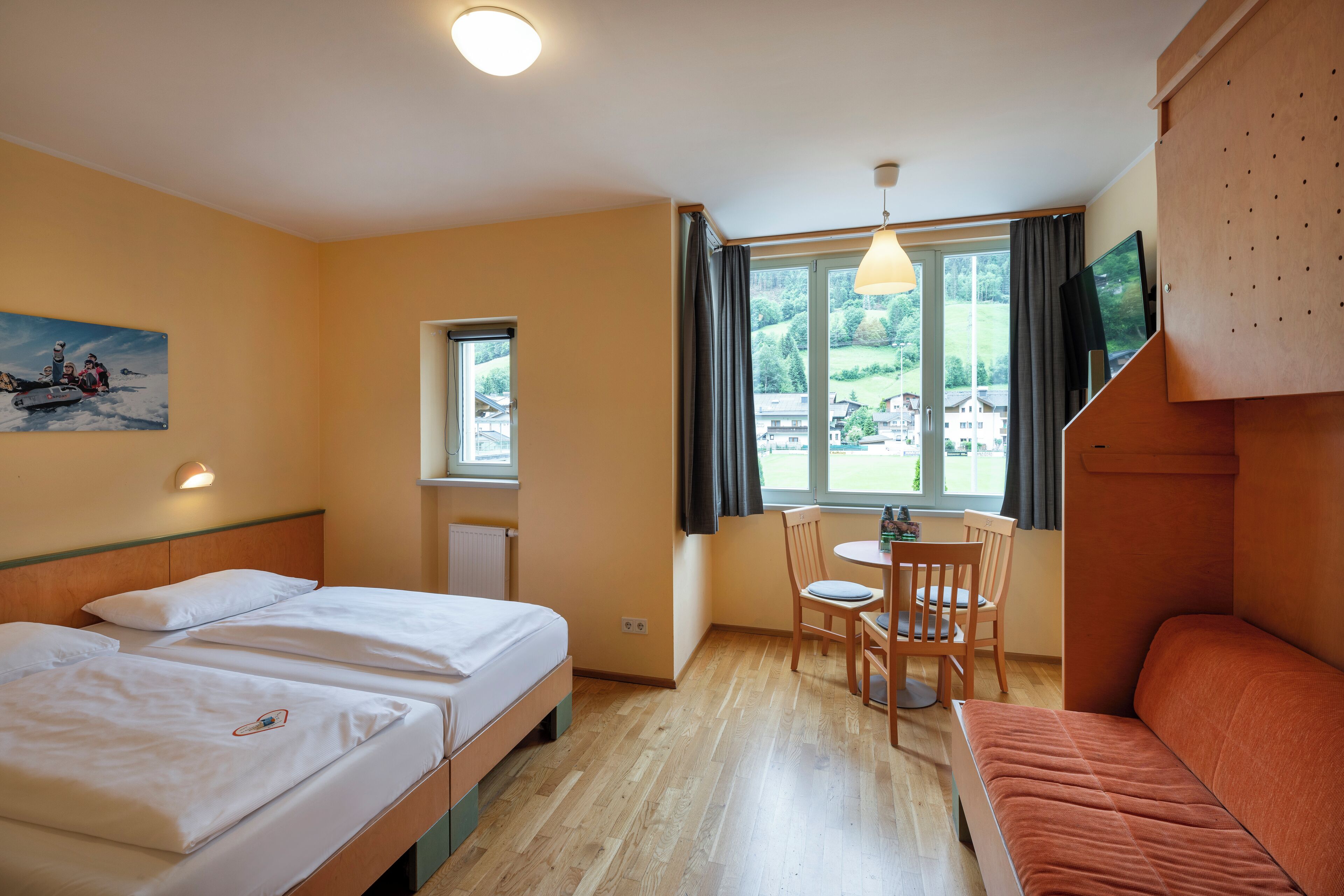 Habitación de hotel con dos camas, una mesa de comedor con sillas, un televisor montado en un mueble de madera y una ventana con vista a zonas verdes y edificios.