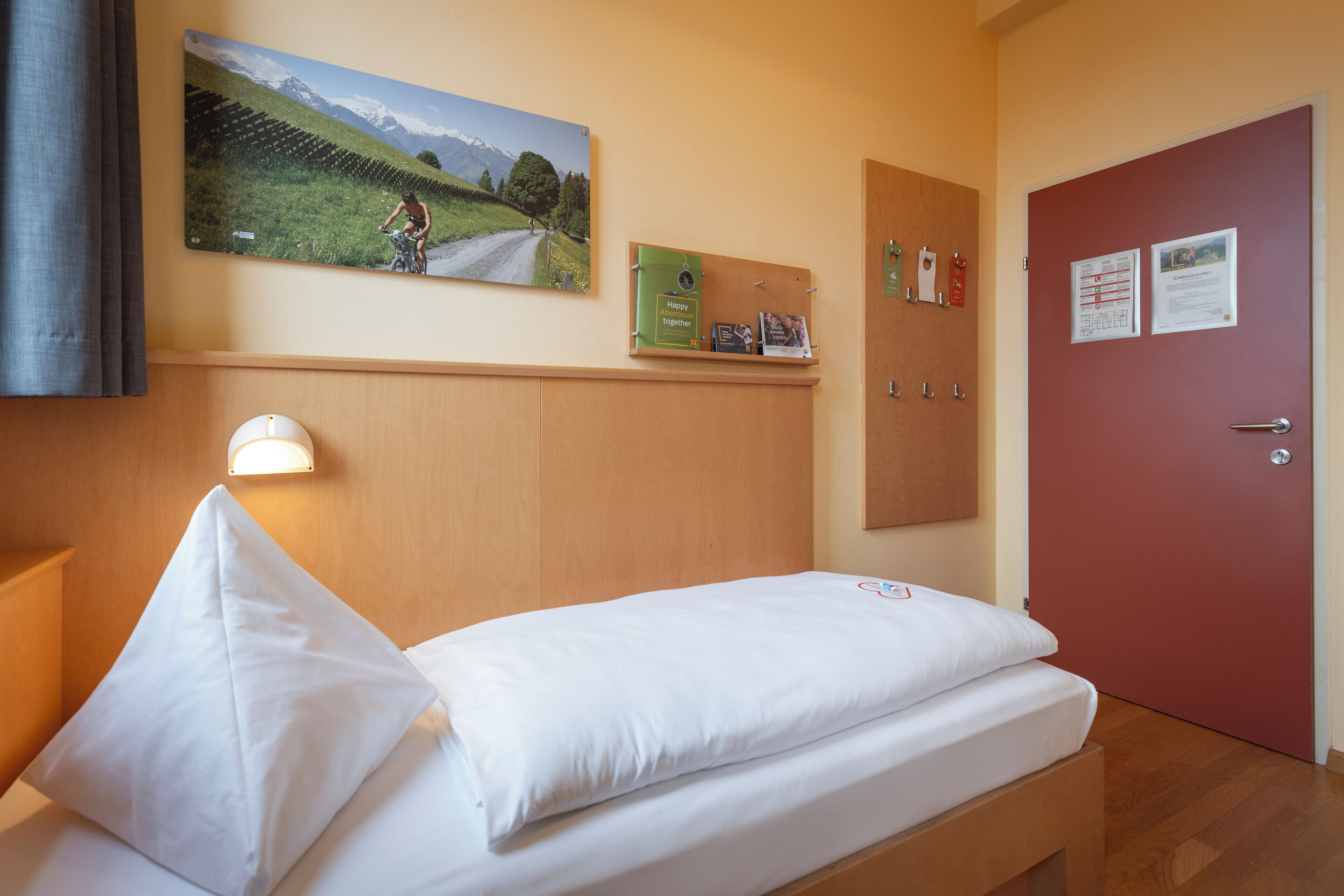A single bed with white bedding, a mounted picture of a cyclist on a road, a bulletin board with a green folder, and a red door with two notices.