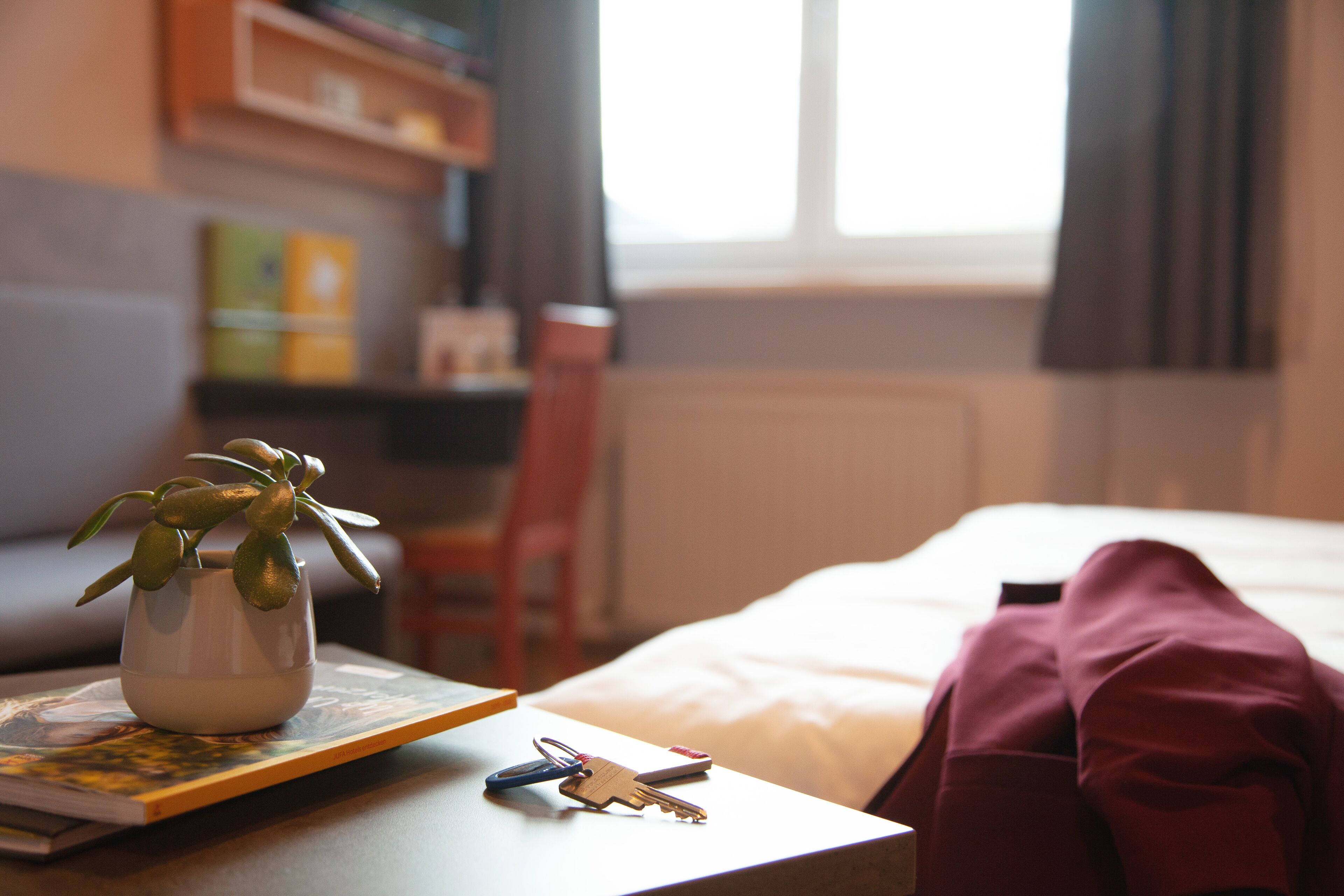 Ein ordentlich eingerichtetes Hotelzimmer mit einem Bett, einem Nachttisch mit Schlüsseln und einer Pflanze, einem Bücherregal und einem Fenster mit Vorhängen.