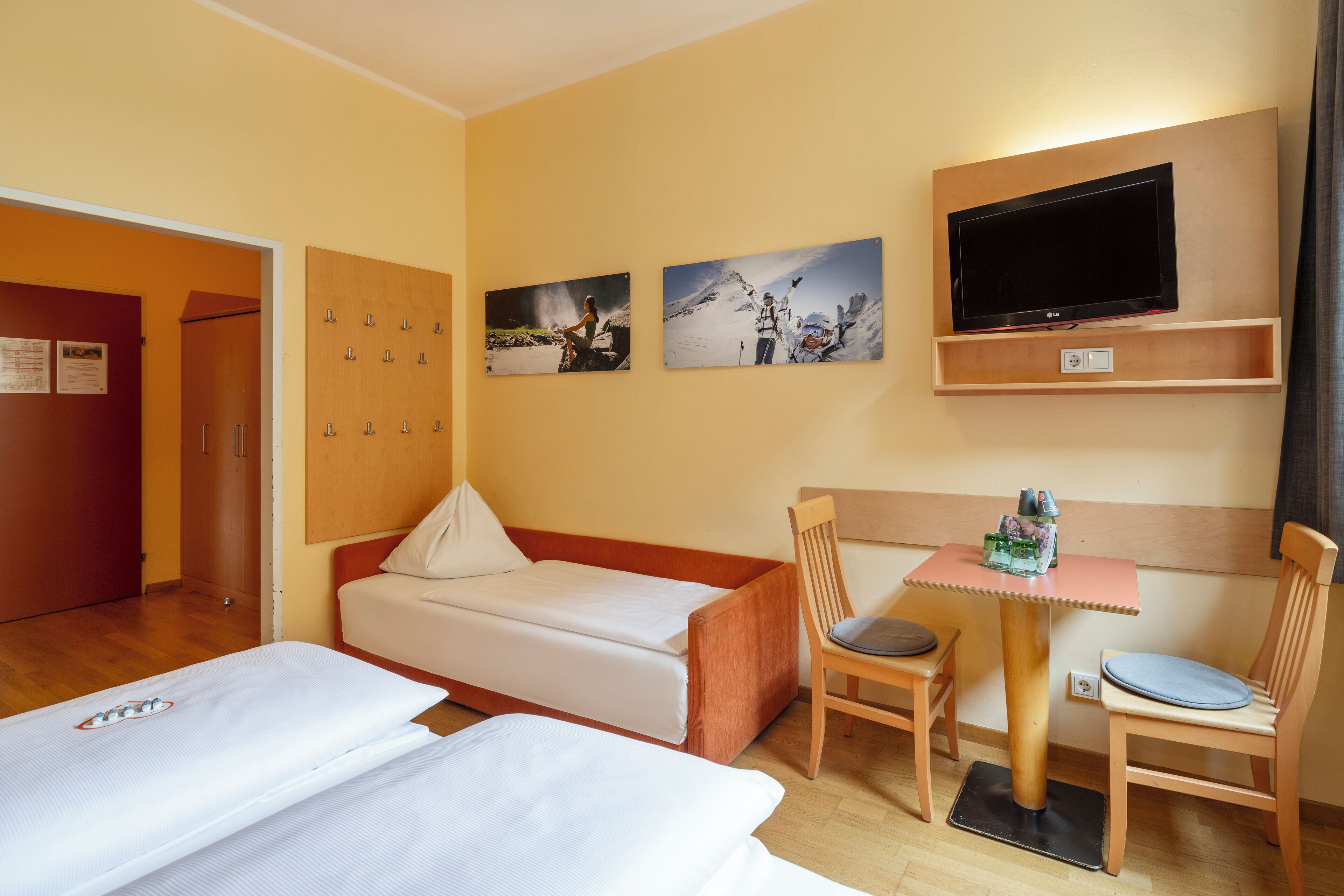 A hotel room with two single beds, a small table with chairs, a TV mounted on the wall, and a wooden cabinet with hooks.