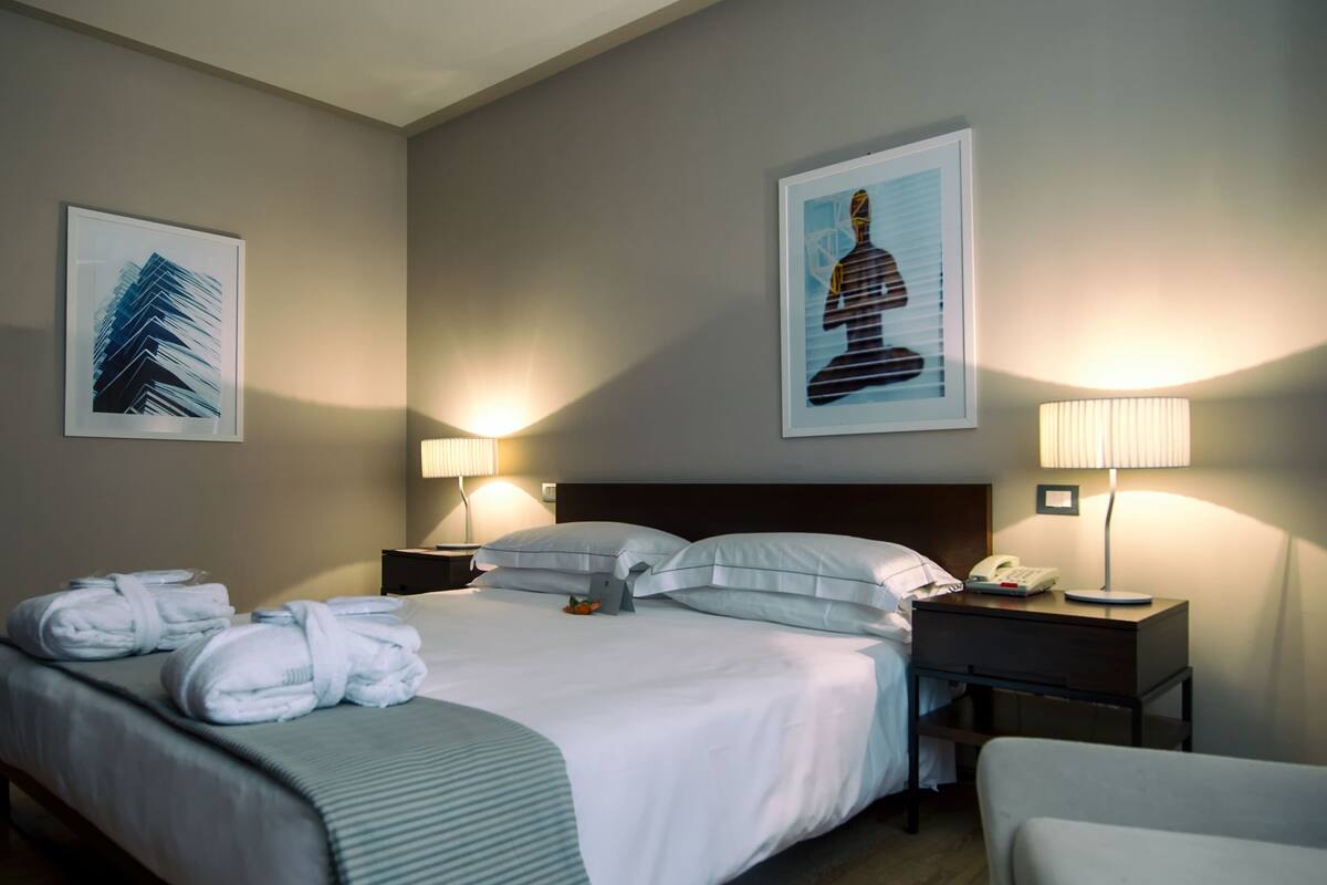 A hotel room with a neatly made bed, two bedside tables with lamps, a chair, and artwork on the walls.