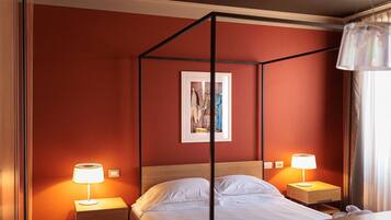 Camera d'albergo con letto a baldacchino, due lampade ai lati del letto, un quadro incorniciato appeso al muro e una finestra con tende.