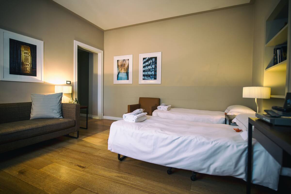 A hotel room with a sofa, two beds, a desk, and wall art.