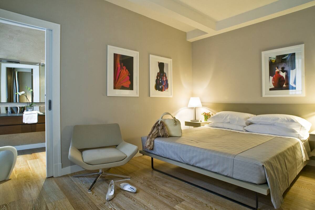 A hotel room with a bed, a chair, a bedside table with a lamp, and artwork on the walls.