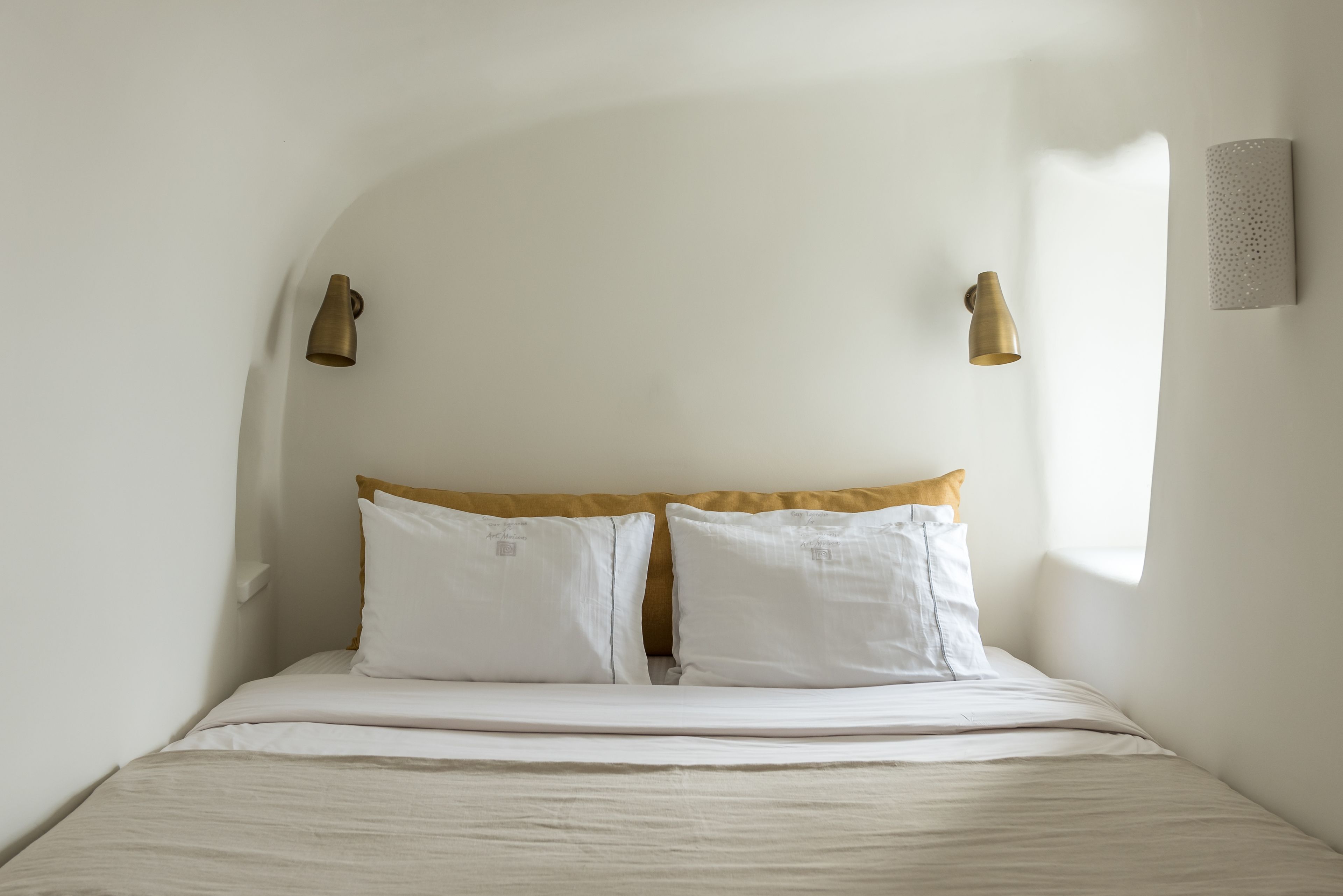 A bed with white bedding and a beige headboard, two wall-mounted light fixtures, and a textured wall panel.