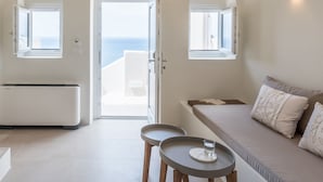 A modern, minimalist room with a large arched ceiling, a central light fixture, two small tables, a sofa with cushions, and a view of the sea through an open door.