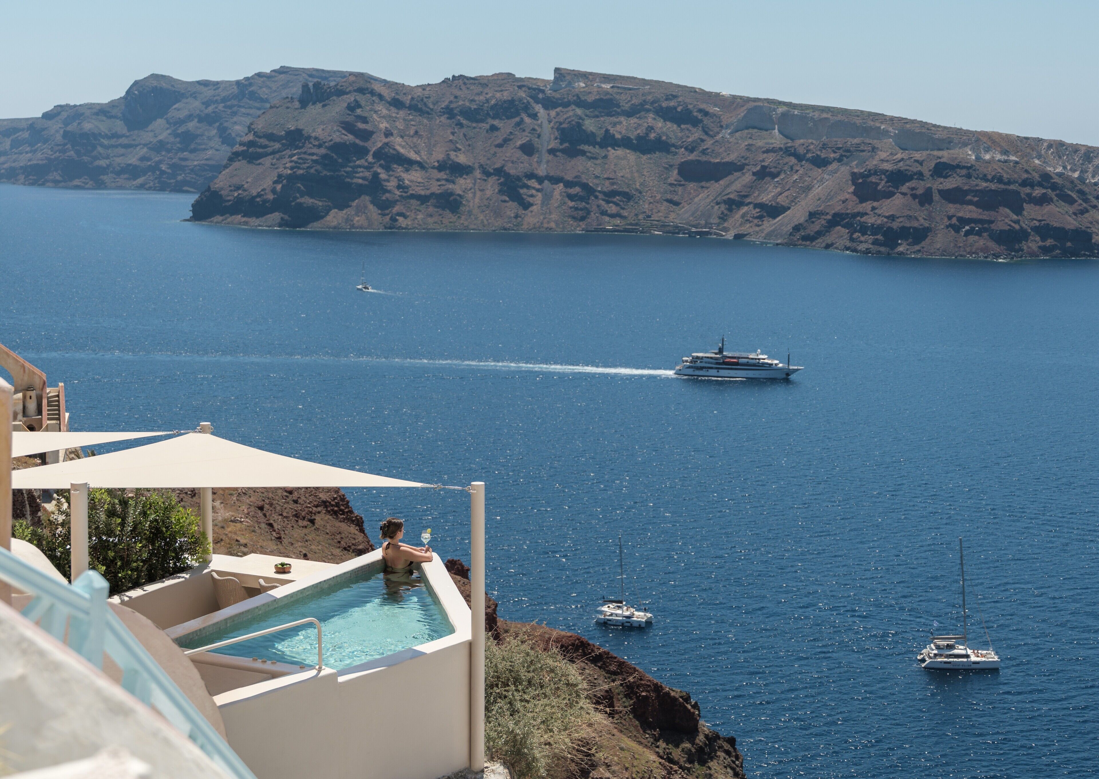 A pool area with a view of the sea, a yacht, and rocky cliffs.