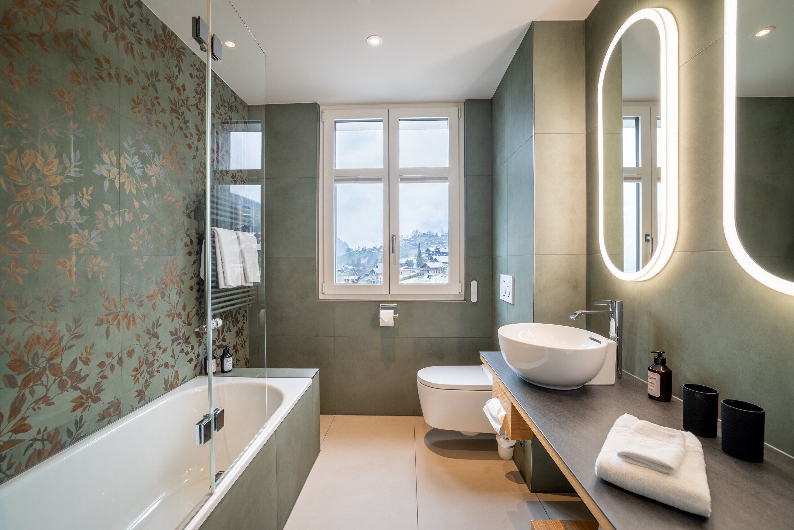 A modern bathroom with a bathtub, toilet, and sink. There is a window providing a view of the cityscape.