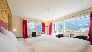 A hotel room with a large bed, a desk with a laptop, and a view of mountains through the windows.