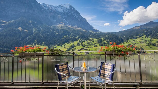 A balcony with two chairs and a table, overlooking a mountainous landscape with a lake and red flowers.