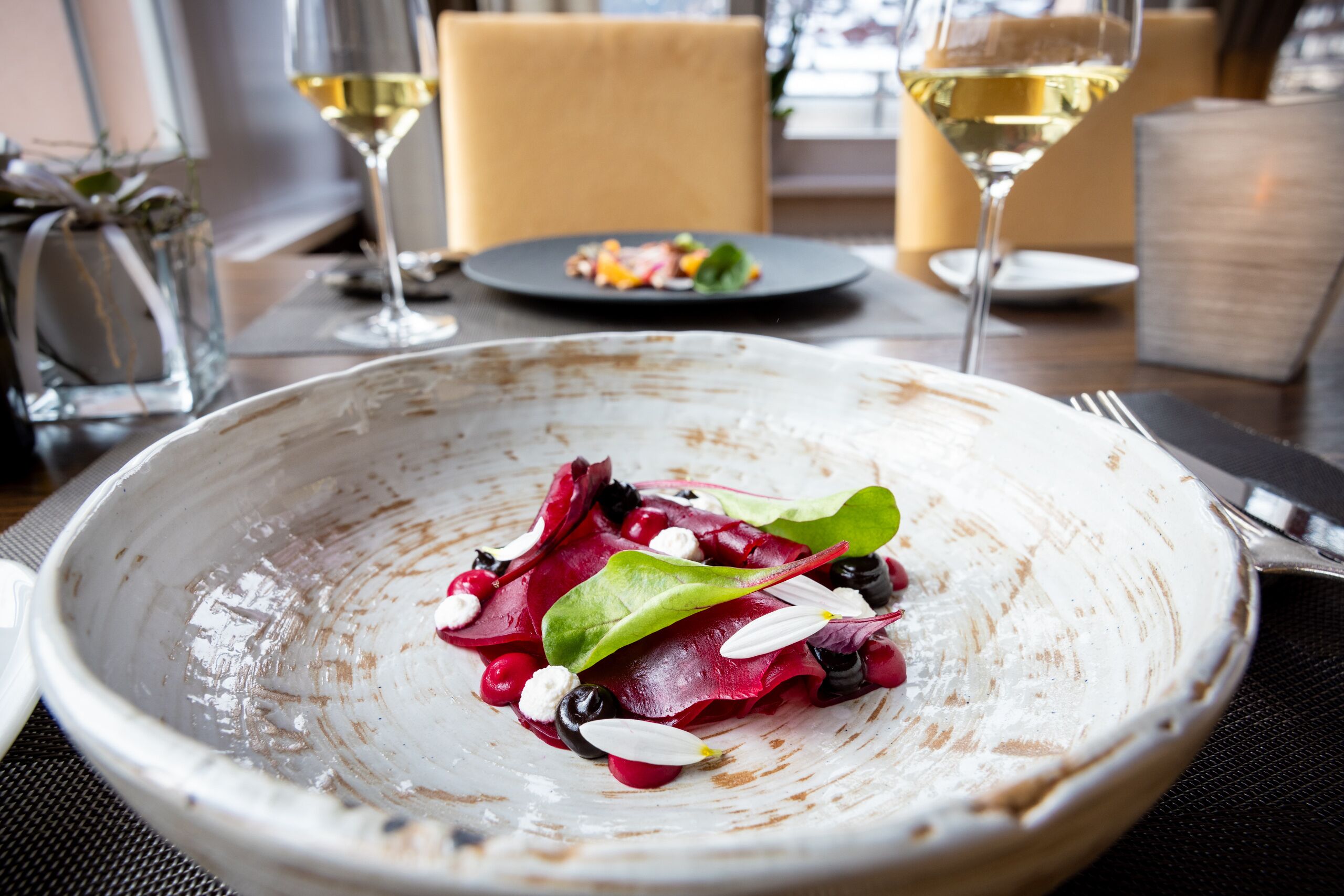 A dining table with a white ceramic bowl containing a salad, two wine glasses, and a plate with a small dish of food.