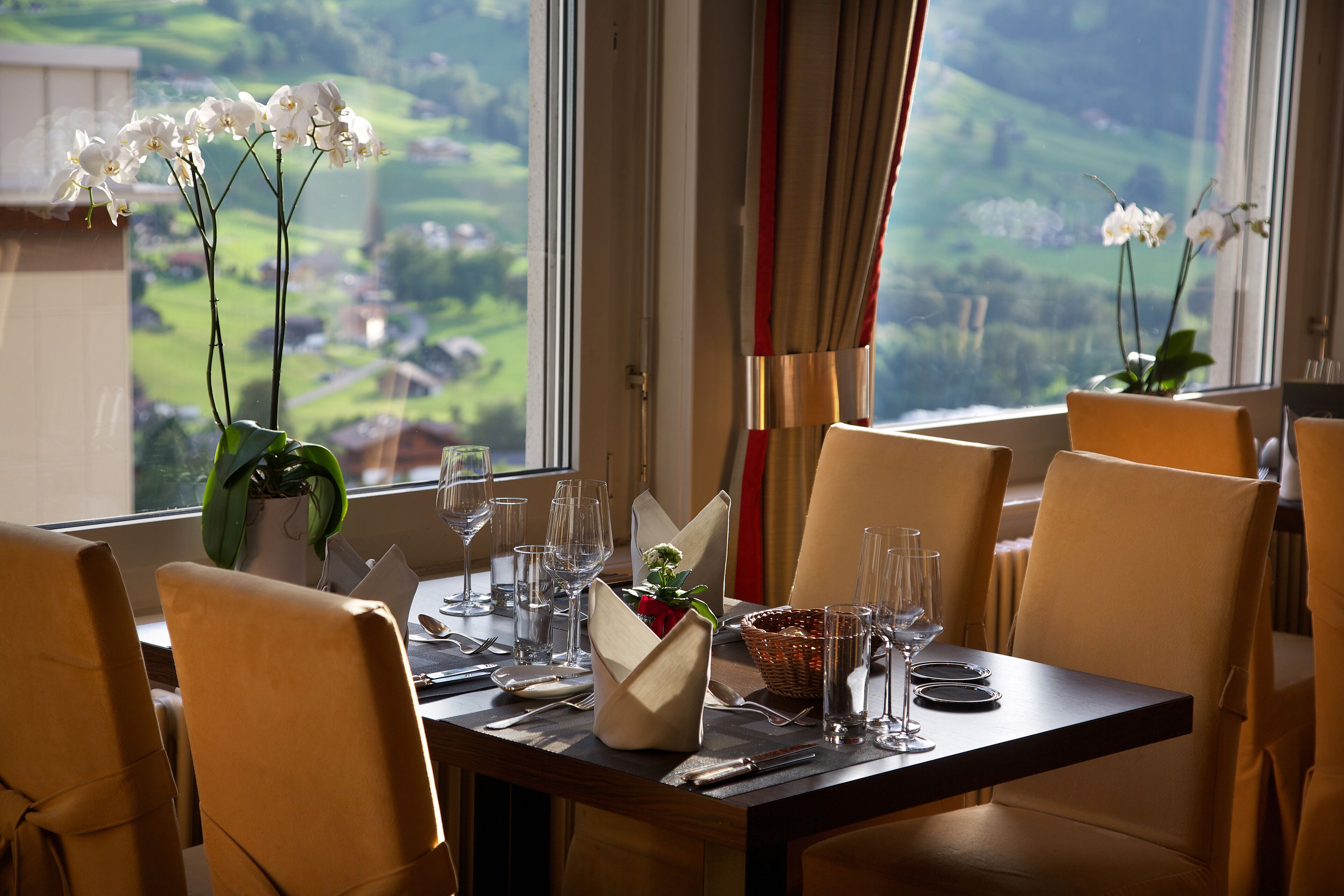 A dining table set for four with chairs, a vase with flowers, and a view of a green landscape through the window.