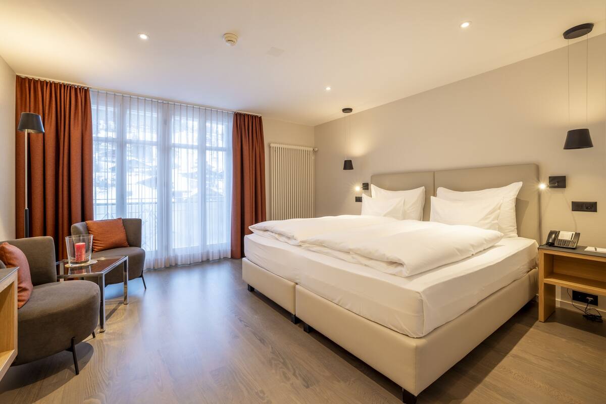 Quarto de hotel moderno com uma cama grande, uma escrivaninha, uma cadeira e vista para a cidade por grandes janelas.