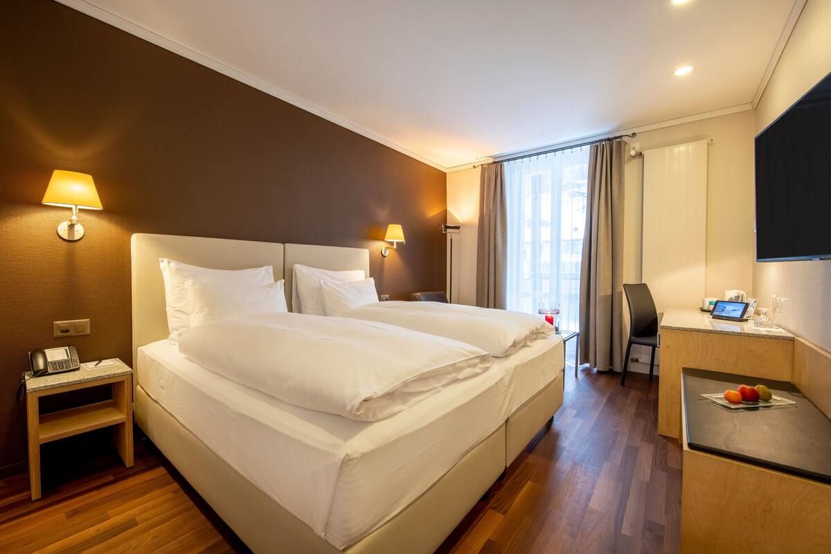 Quarto de hotel com uma cama grande, luminárias de cabeceira, uma escrivaninha com computador e uma televisão.