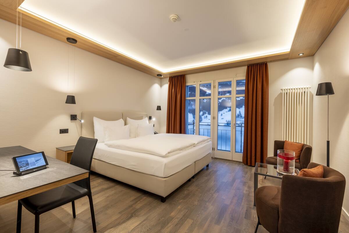 Quarto de hotel moderno com uma cama, uma escrivaninha com um laptop, uma cadeira e vista para uma paisagem nevada pela janela.