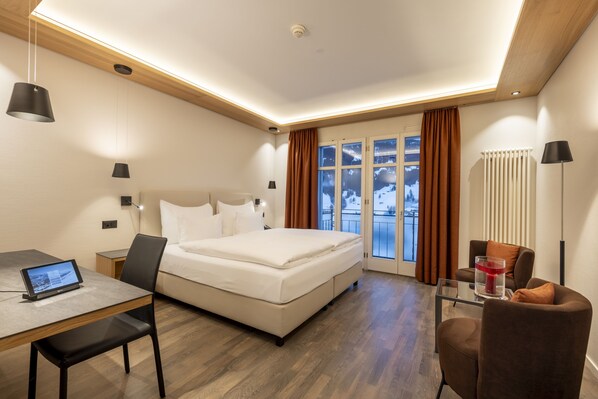 A modern hotel room with a bed, a desk with a laptop, a chair, and a view of a snowy landscape through the window. - Belvedere Swiss Quality Hotel (Grindelwald)