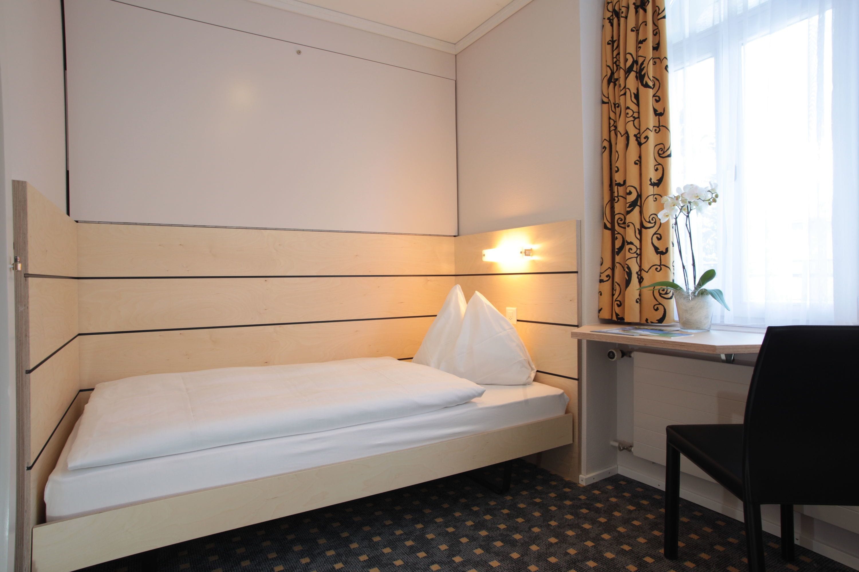A single bed with a headboard, a desk with a chair, a window with curtains, and a potted plant.