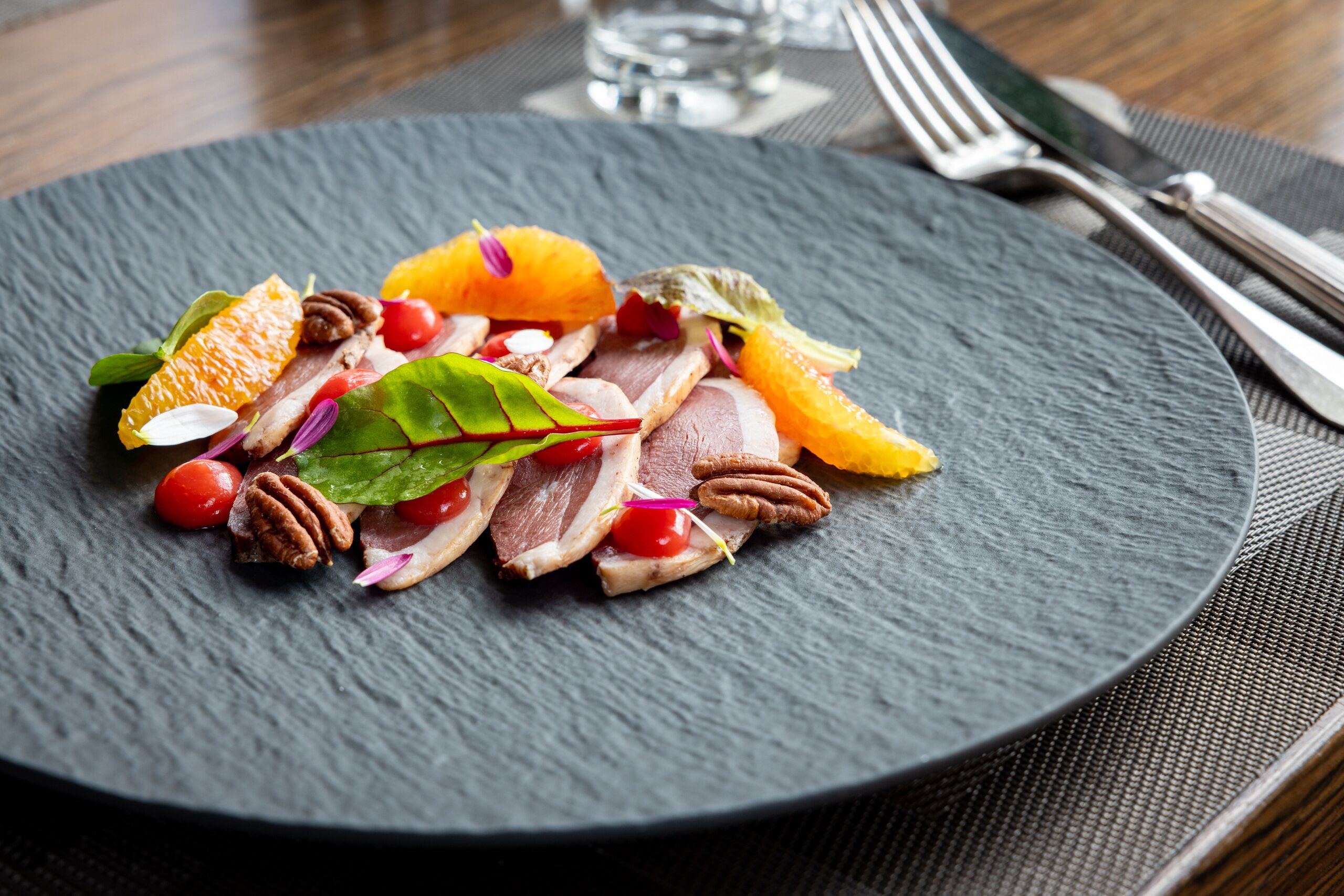 Uma travessa de carne fatiada com frutas e nozes sobre um prato de ardósia escura.