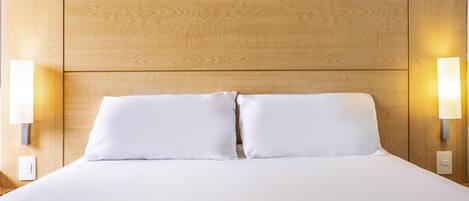 A hotel room with a wooden headboard, two white pillows, and wall-mounted lights.