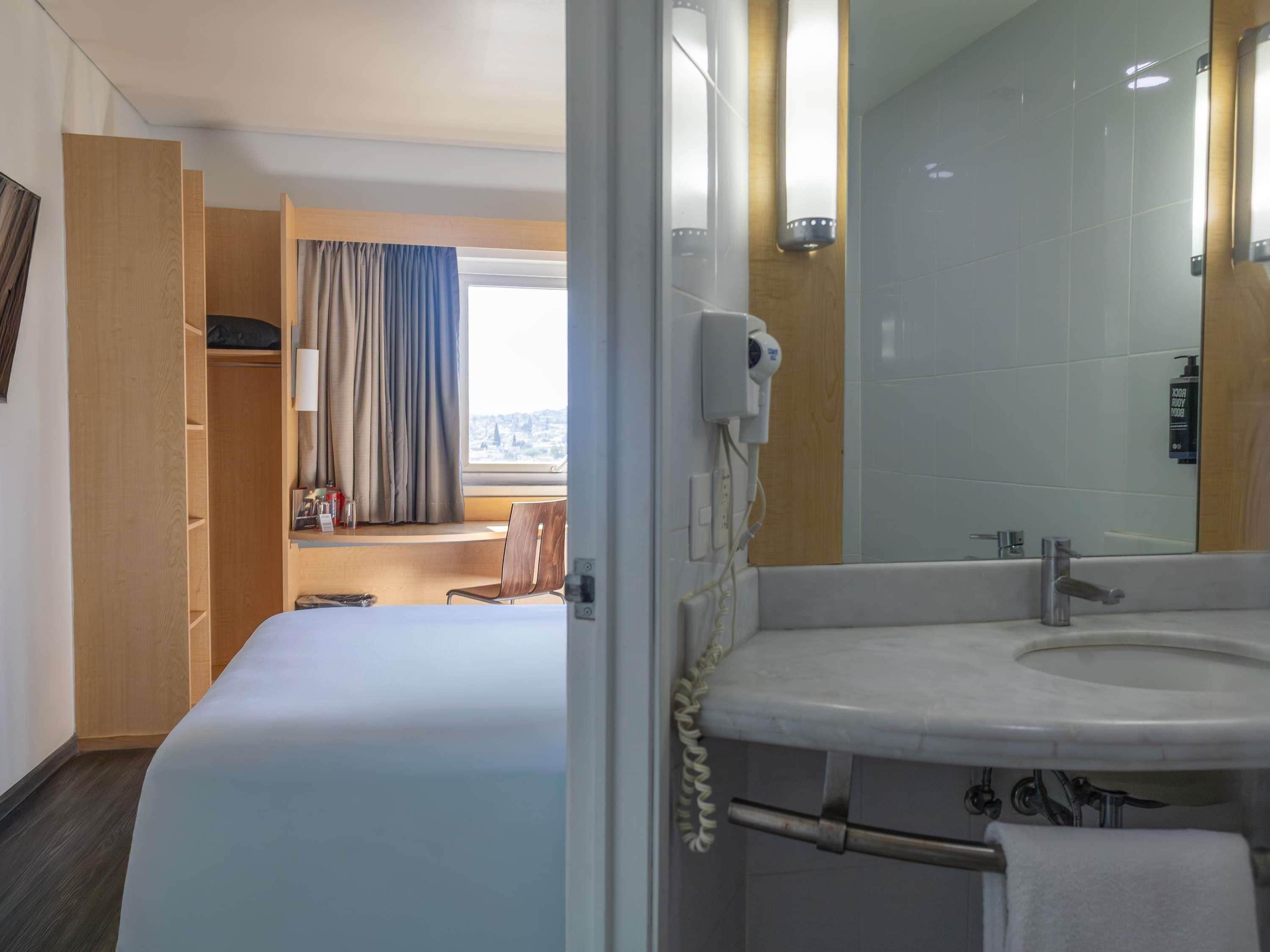 Et hotelværelse med seng, badeværelse med håndvask og spejl, samt udsigt over byen fra vinduet.