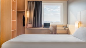 A hotel room with a bed, a desk with a laptop, a chair, a window with curtains, and a wardrobe.