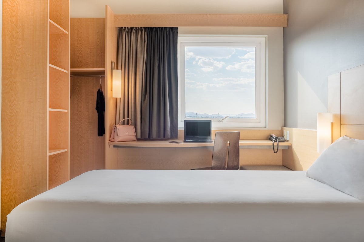 Ett hotellrum med en säng, ett skrivbord med en bärbar dator, en stol, ett fönster med gardiner och en garderob.