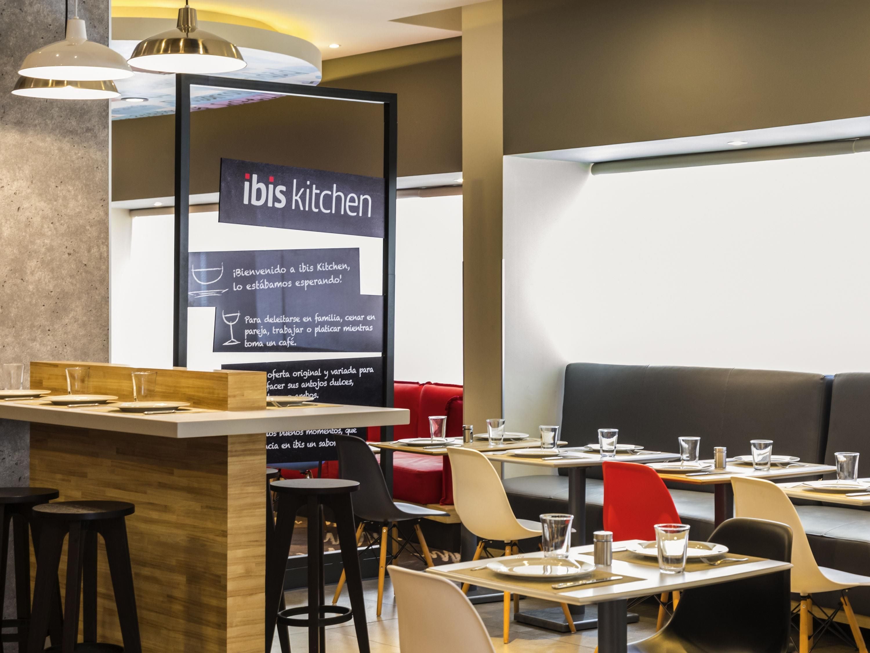 Interior moderno de un restaurante con un letrero que dice 'ibis kitchen' y una pizarra de menú. Las mesas están dispuestas con platos y vasos.