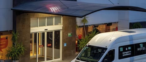 Ein Hotel Eingang mit einem auffälligen roten Logo, einem davor geparkten Wagen und freiem Blick auf die Eingangstüren.