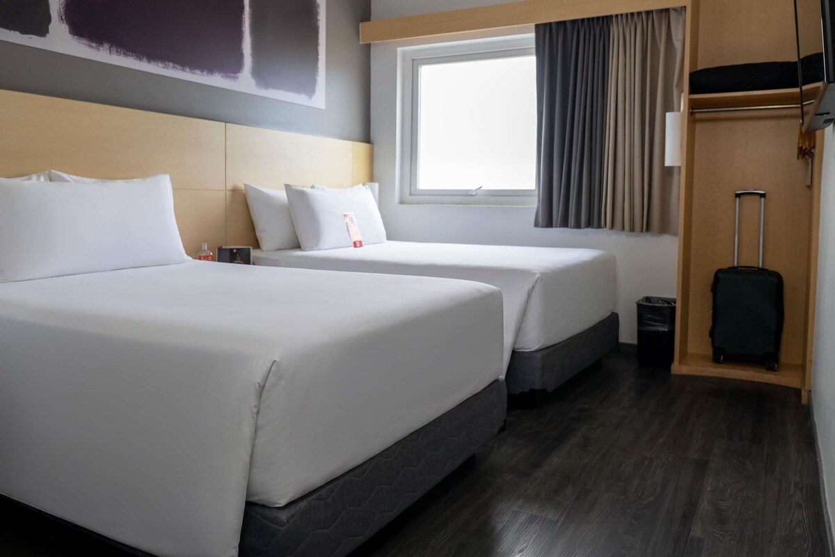 Habitación de hotel con dos camas, una ventana con cortinas, un cabecero de madera y una maleta.