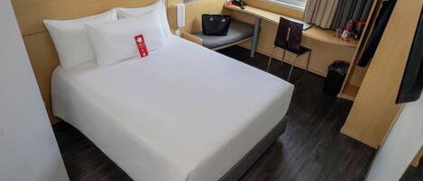 Habitación de hotel con cama, escritorio, silla, mesita, cubo de basura y ventana con cortinas.