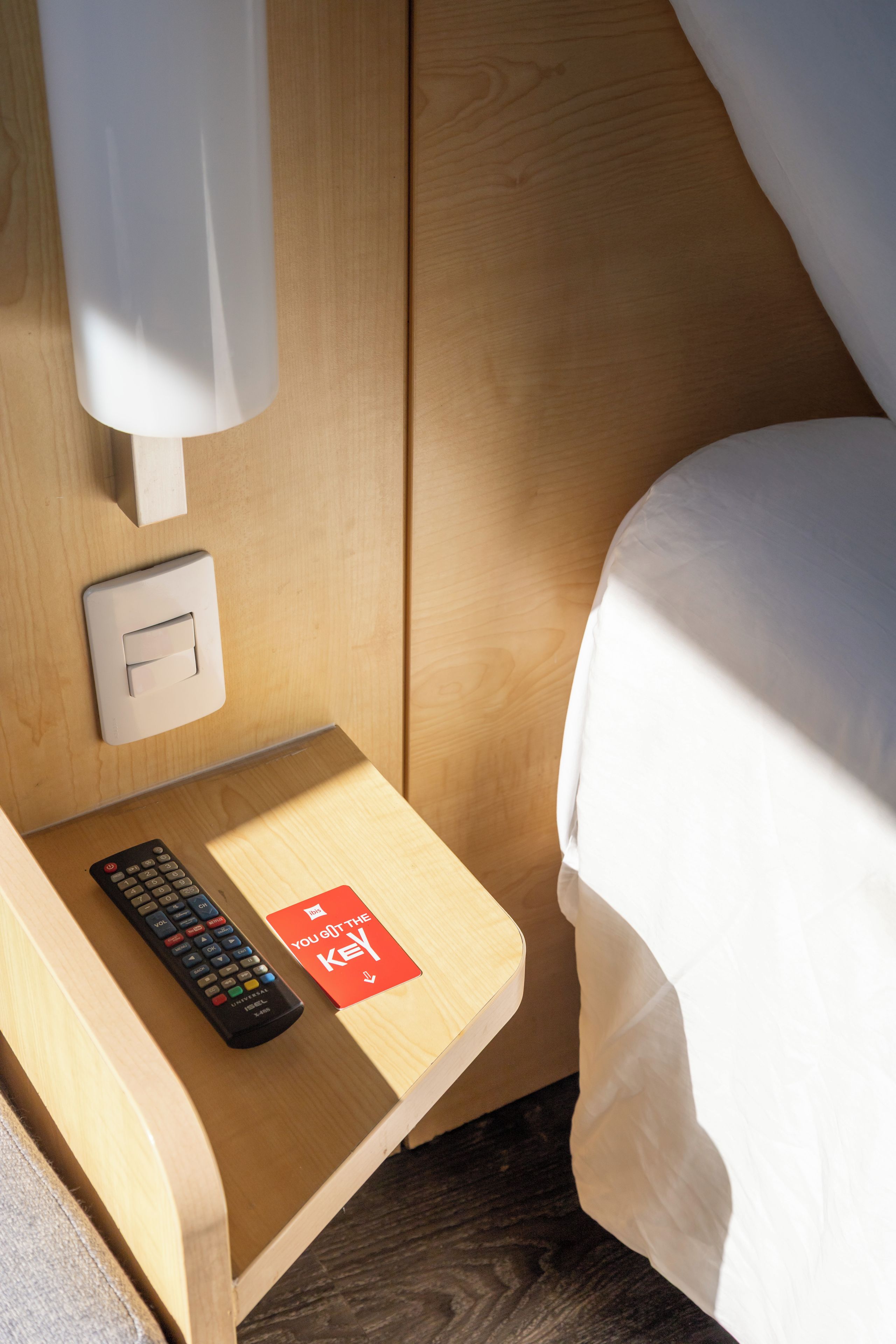 Una mesita de noche de madera con un control remoto y una tarjeta que dice 'You Got the Key', un interruptor de luz y una cama con ropa blanca.