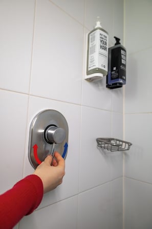 En hand som vrider på en spak på en väggmonterad kontrollpanel, med två flaskor schampo och ett handdukställ av metall.