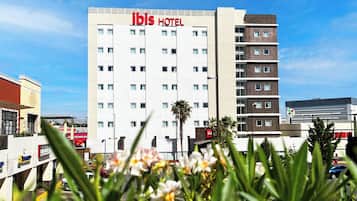 A modern hotel building with a prominent "ibis HOTEL" sign, multiple floors, and a clear blue sky in the background.