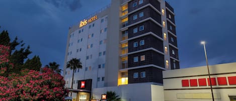Un hotel de varios pisos con un letrero llamativo que dice 'ibis HOTEL', un estacionamiento y una farola.