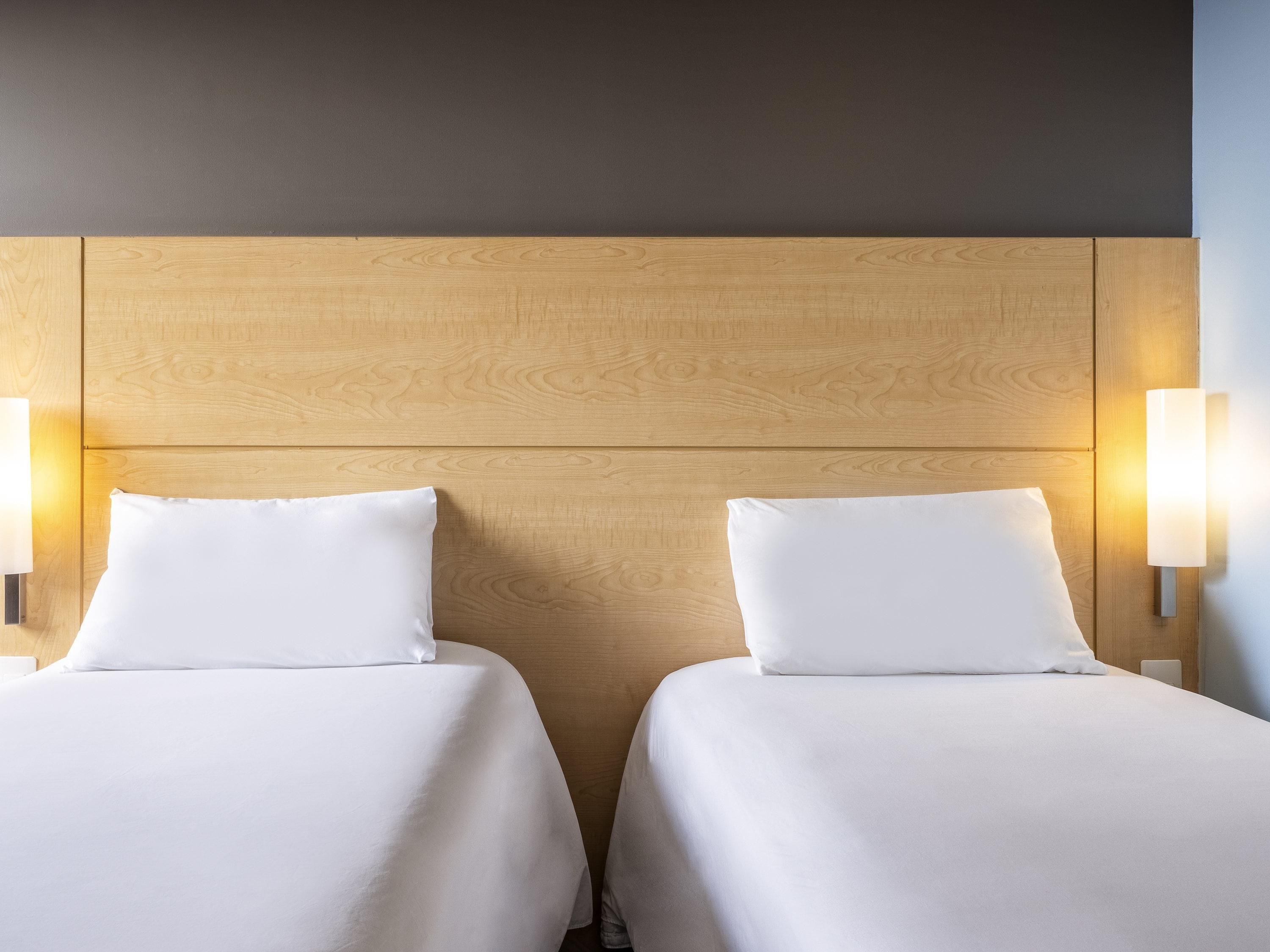 A hotel room with two beds, wooden headboard, and wall-mounted lights.