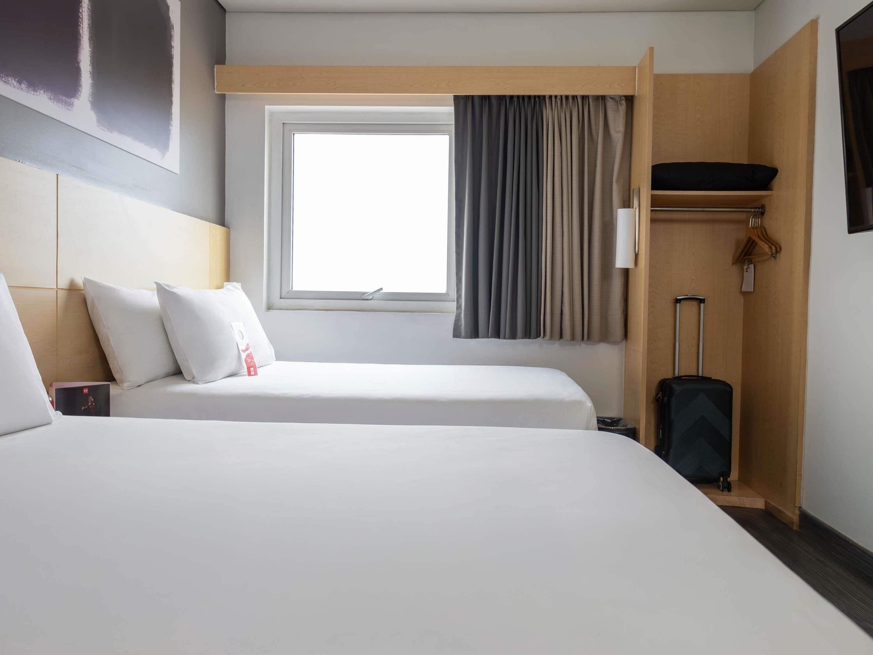 A hotel room with two beds, a window with curtains, a wooden shelf, and a suitcase.