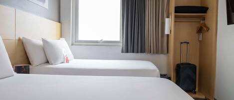 A hotel room with two beds, a window with curtains, a wooden shelf, and a suitcase.
