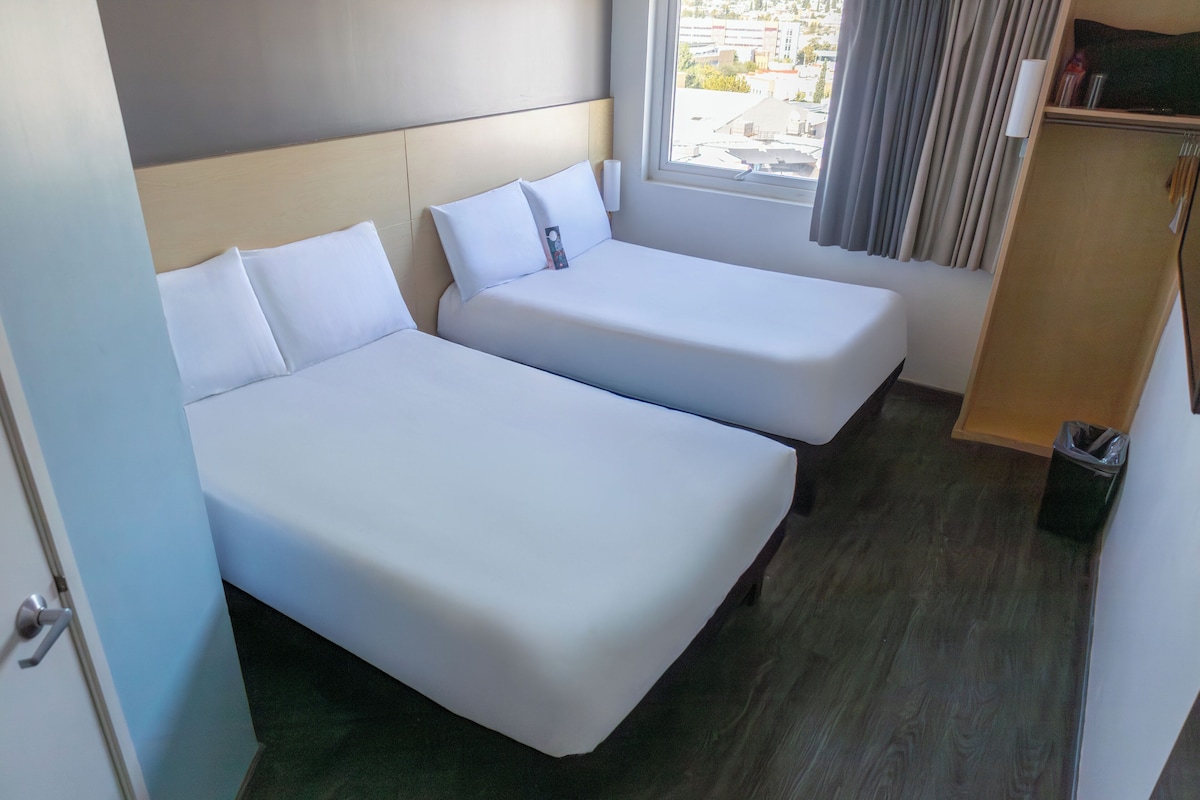 Ett hotellrum med två separata sängar, en träpanel som huvudgavel, ett fönster med gardiner och en liten trägarderob.