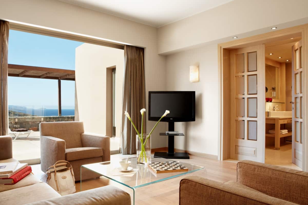 A modern hotel room with a sofa, armchair, and a glass coffee table. There's a TV on a stand, a vase with flowers, and a view of the sea through large windows.