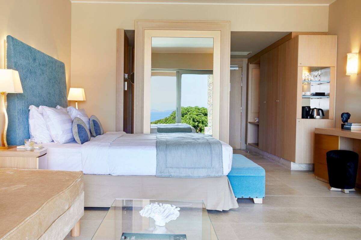 A modern hotel room with a large bed, a sofa, a glass coffee table, and a view of the outdoors.
